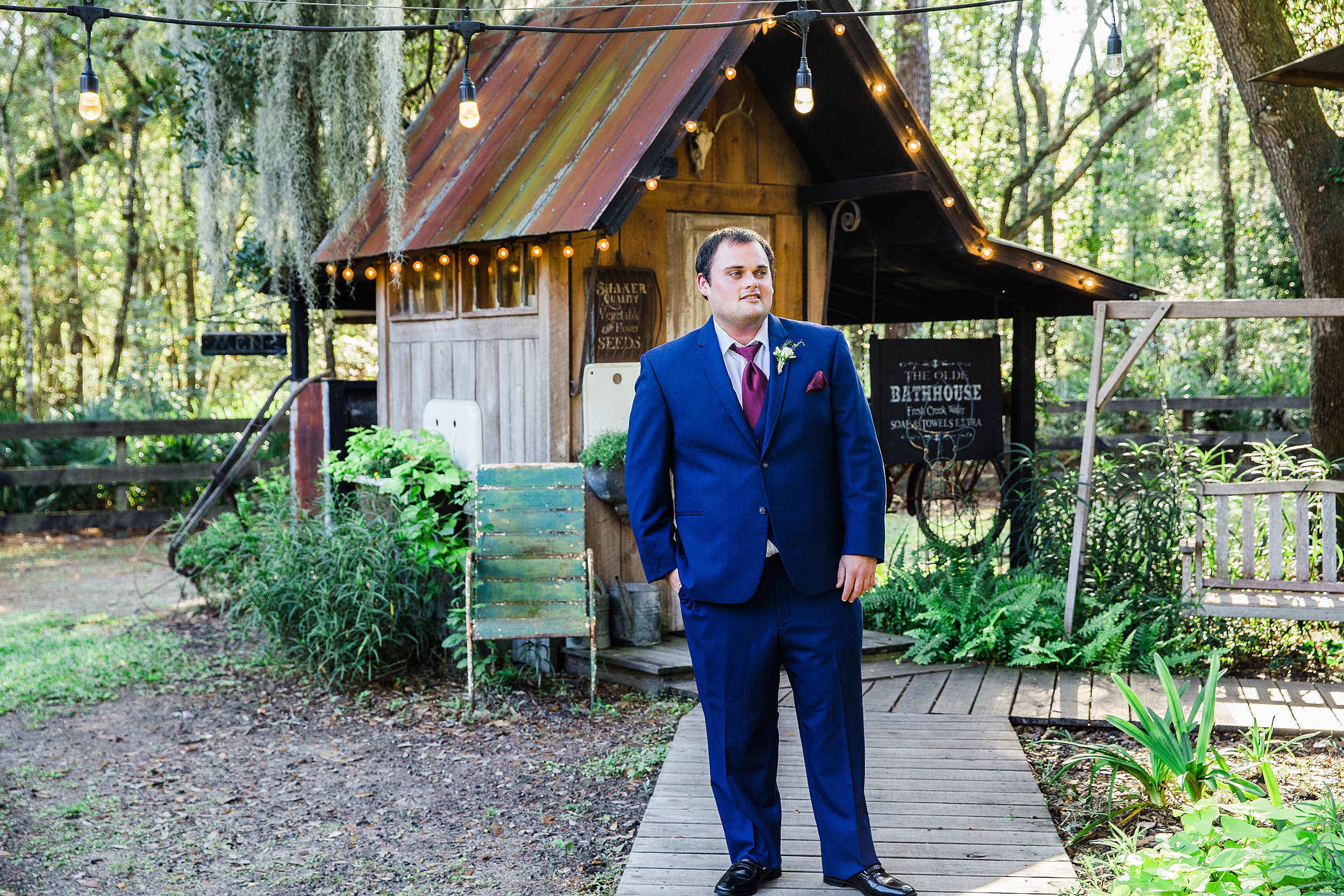 A groom stands in front of an old bathhouse under an oak canopy in a blue suit and maroon details