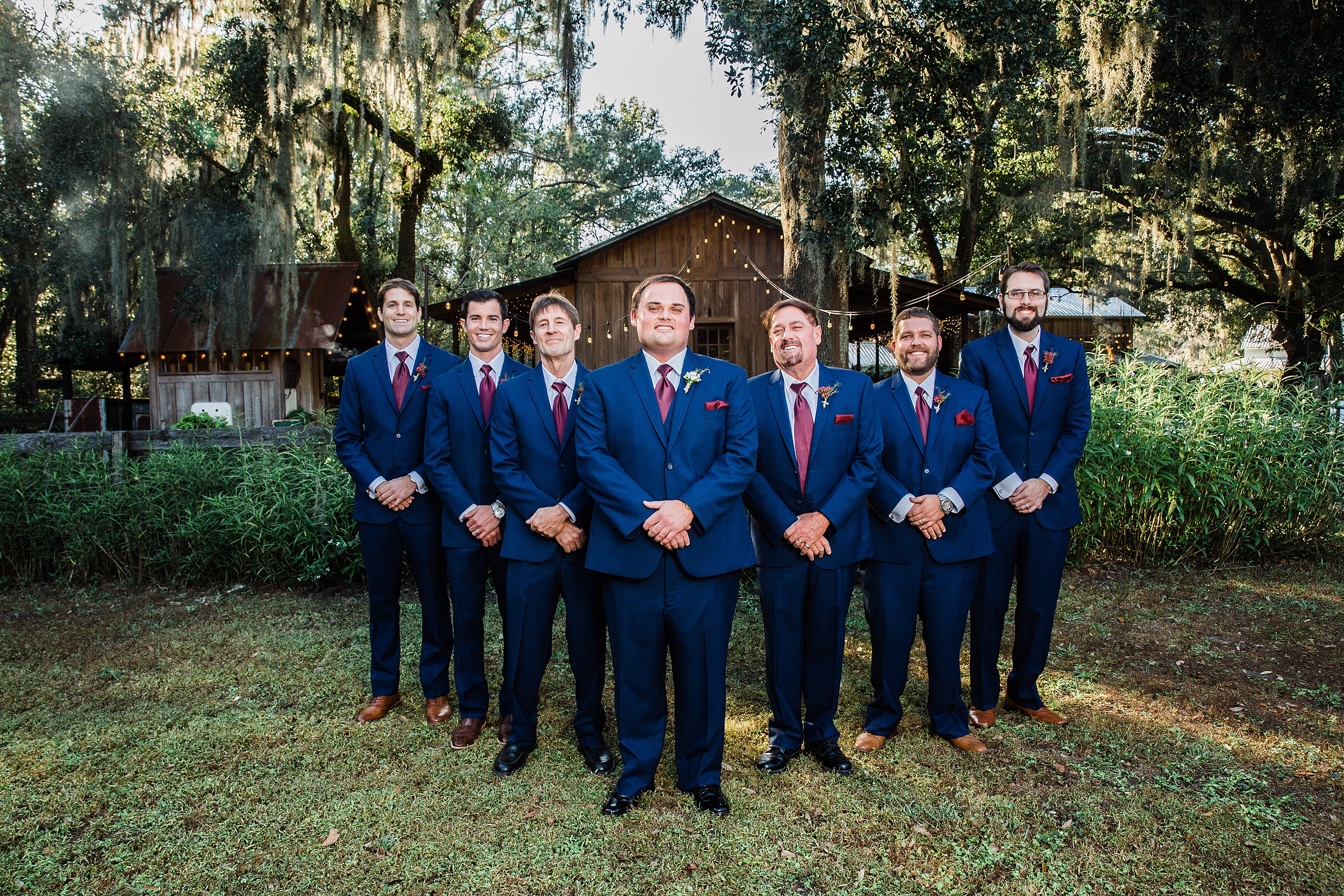 A groom in a blue suit stands with hands crossed with his 6 groomsmen under an oak canopy Tucker's Farmhouse Wedding