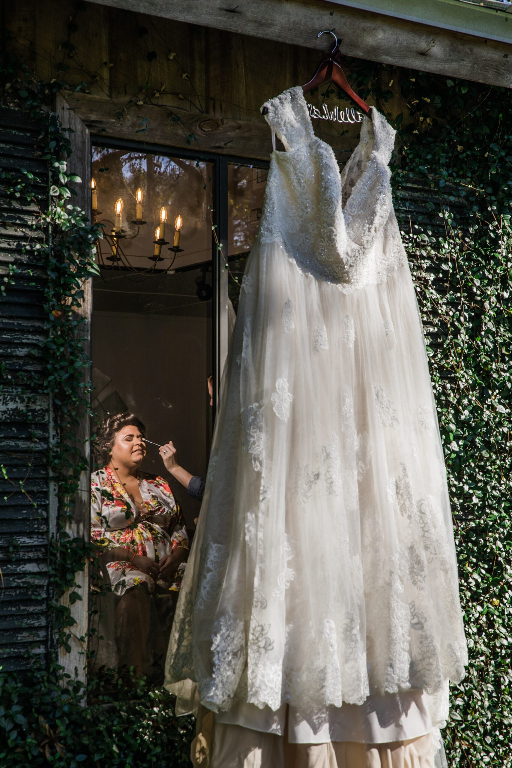 A dress hangs on a vine covered building while the bride gets makeup done inside