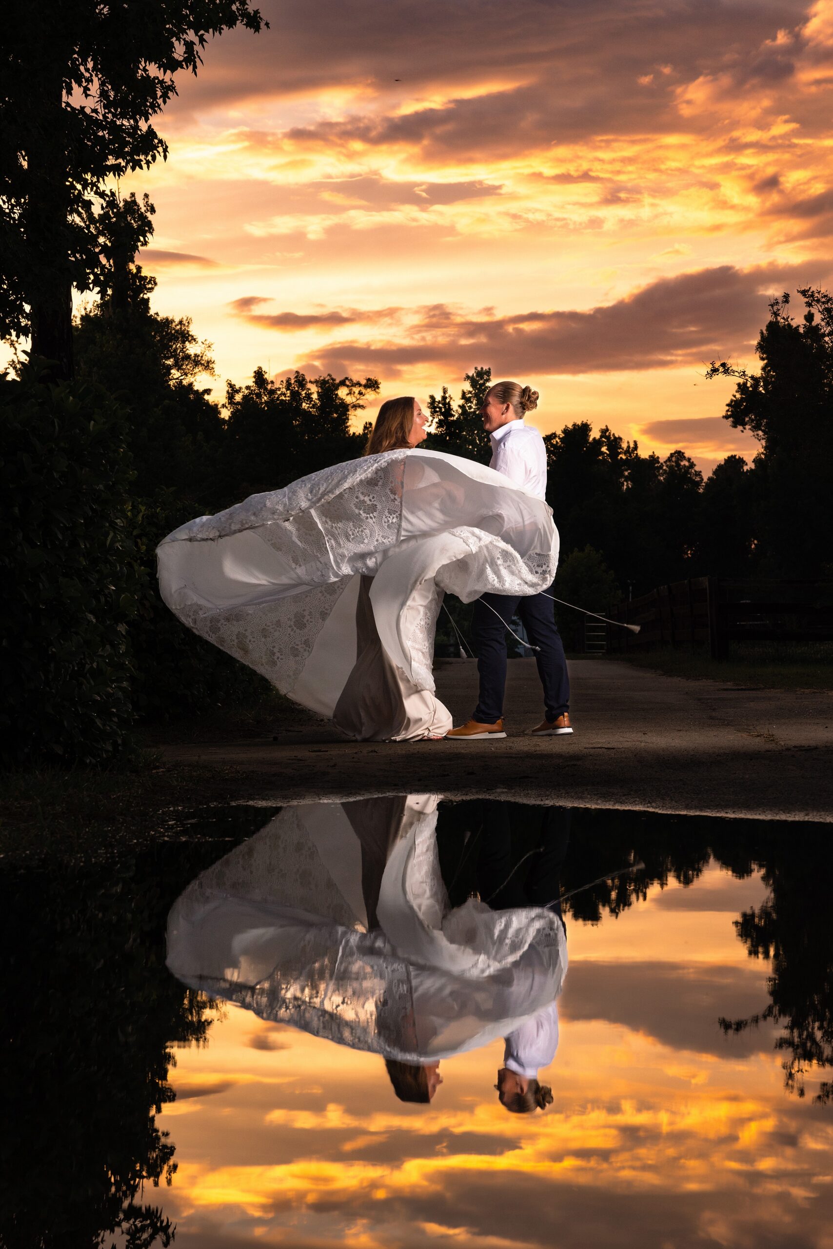 A brides dress flows in the wind while standing in a garden and reflecting in a pond at sunset with her wife at their marsh landing country club wedding