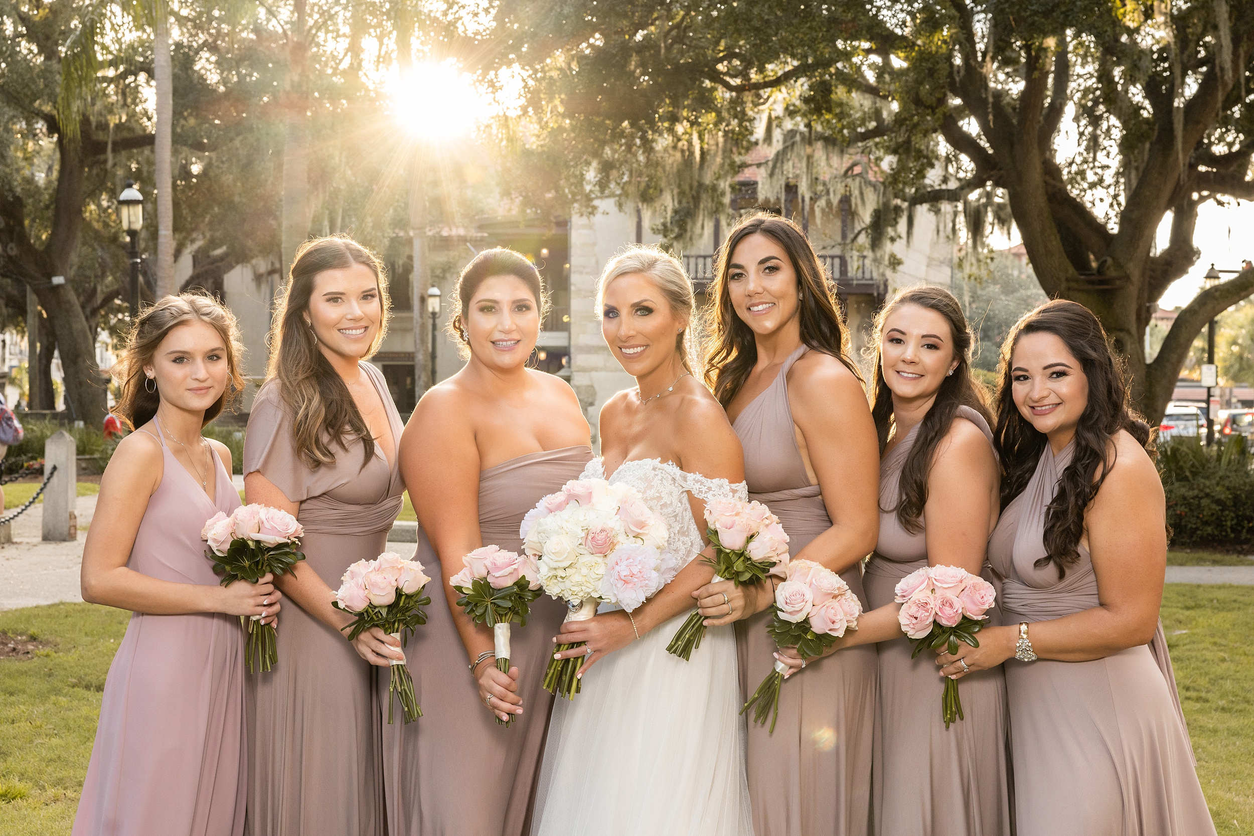 A bride stands with her bridal party at sunset holding their pink rose bouquets