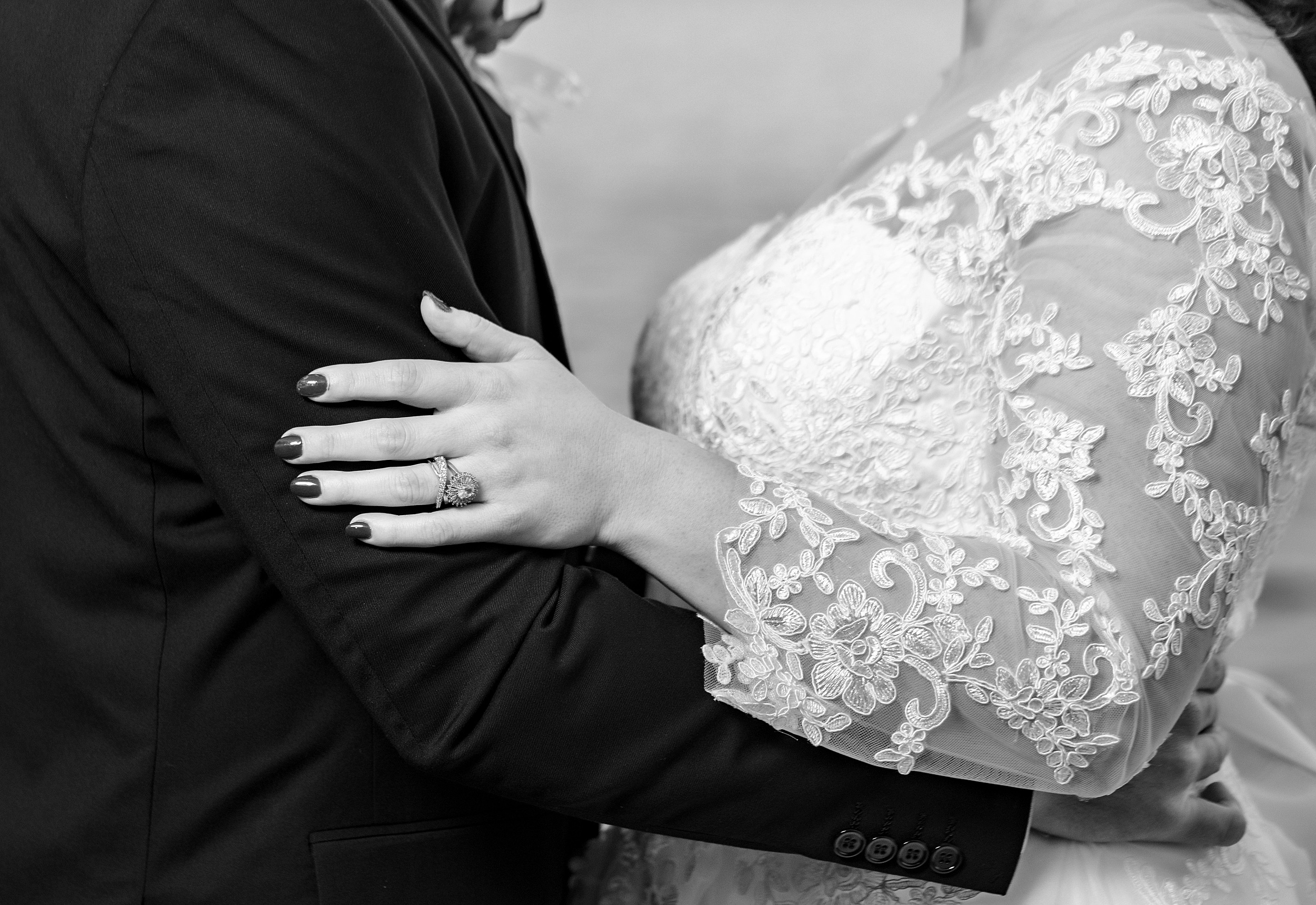 Details of a bride and groom holding each other in a lace embroidered dress and black suit