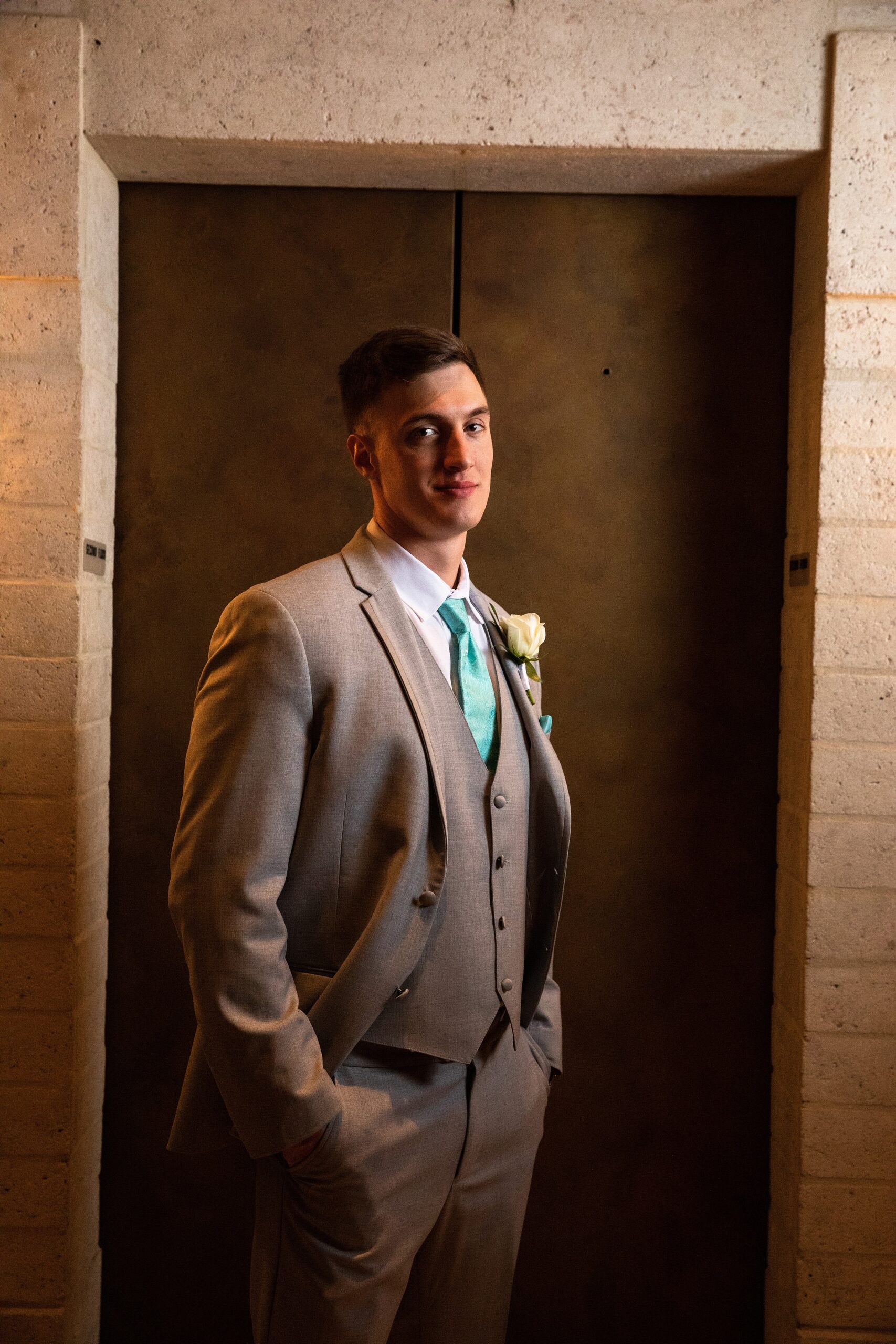 A groom stands in a grey suit and teal tie in front of an elevator door