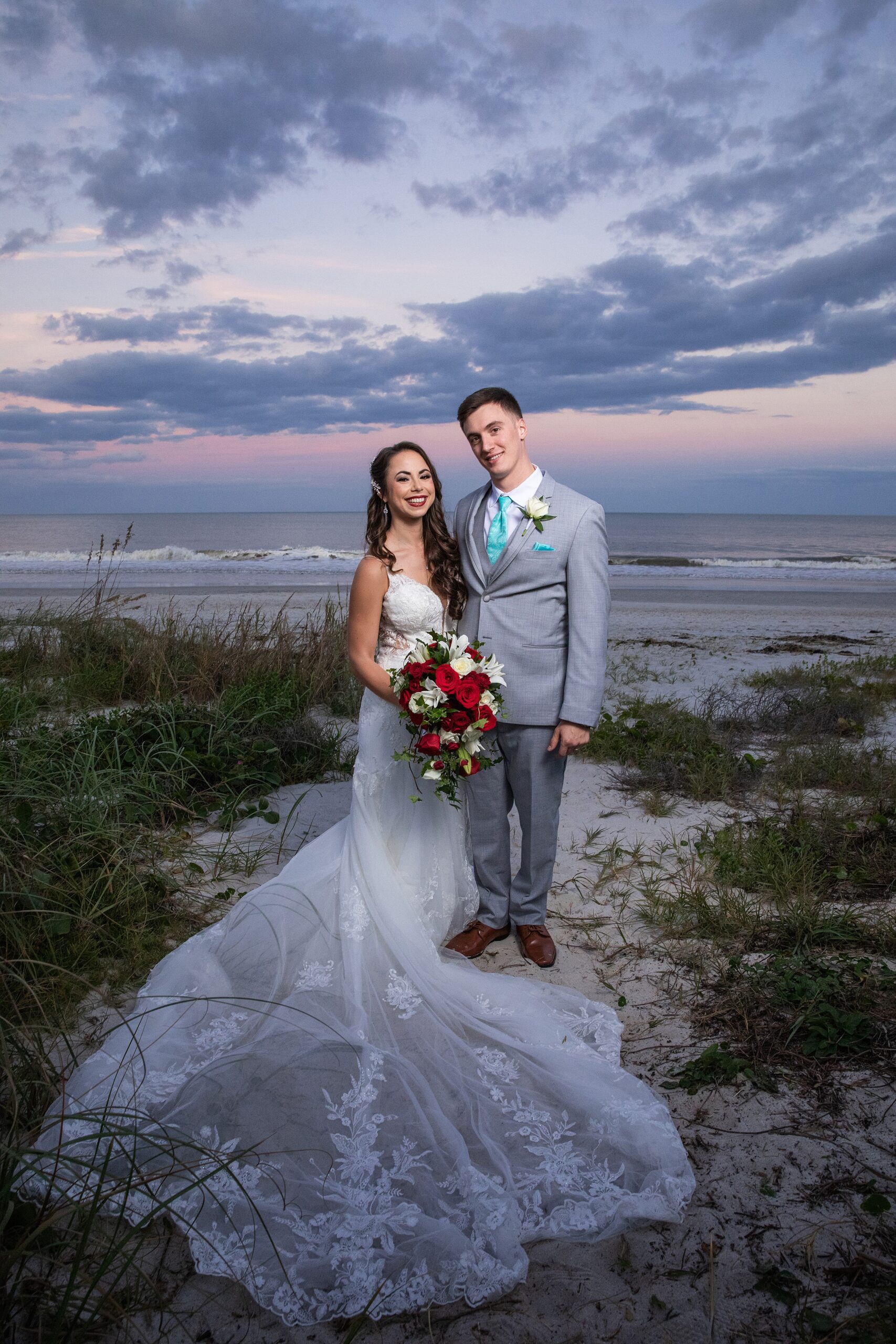 Newlyweds stand in the dunes of the lodge and club ponte verda wedding venue holding each other and the large red and white rose bouquet