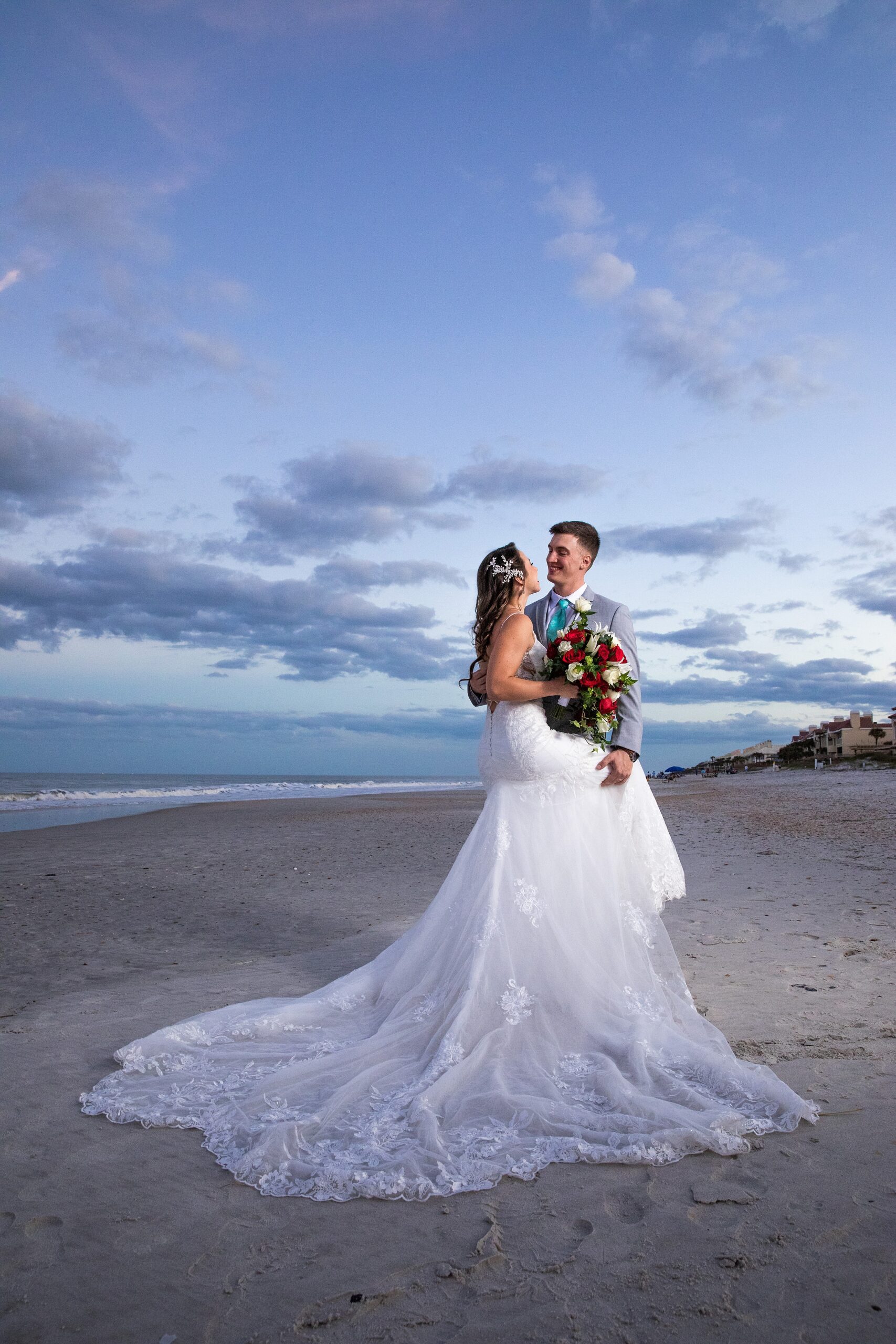 A bride and groom embrace while standing on the beach at sunset