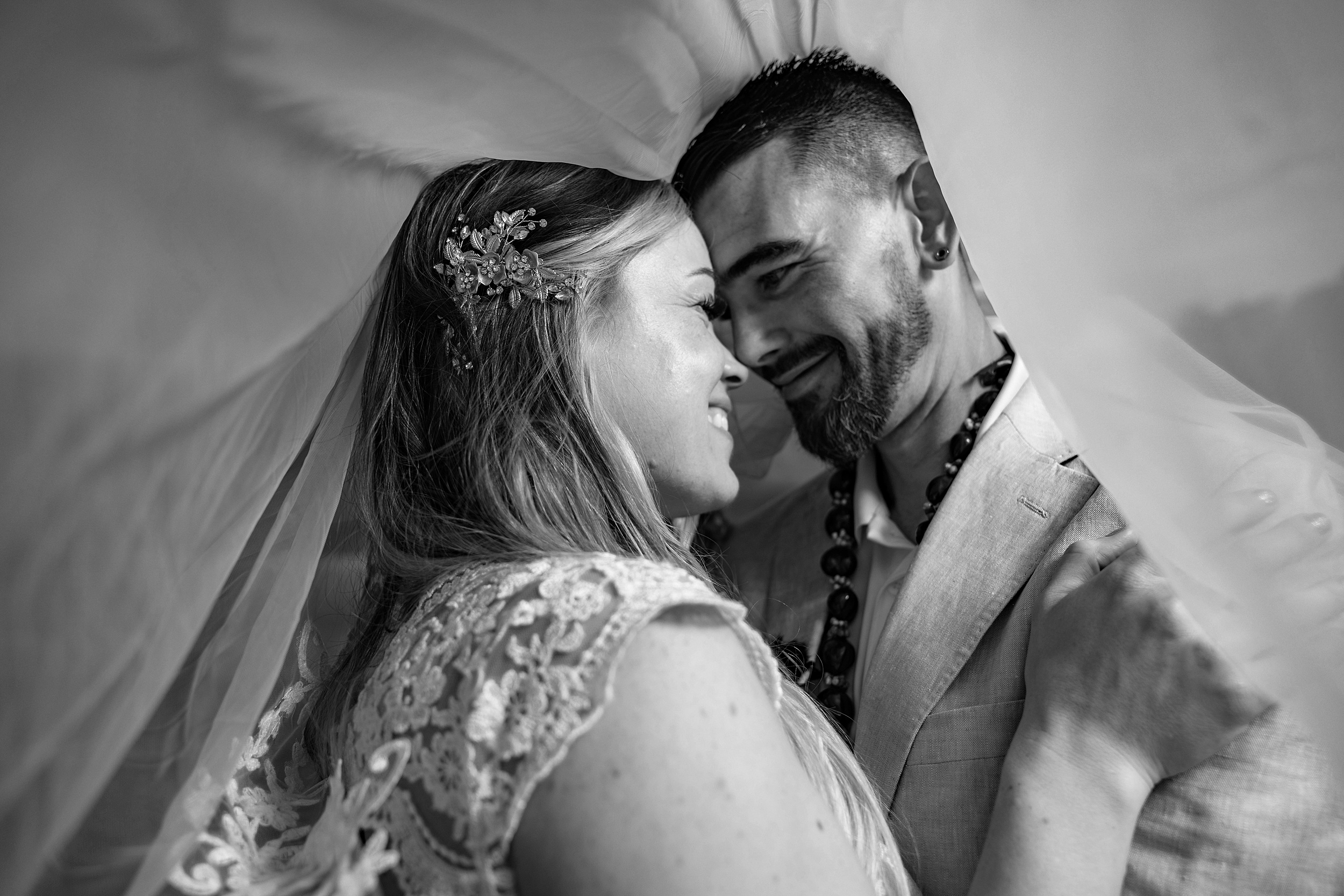 Newlyweds share a smile while standing under a veil in a lace dress at their the garden villa wedding