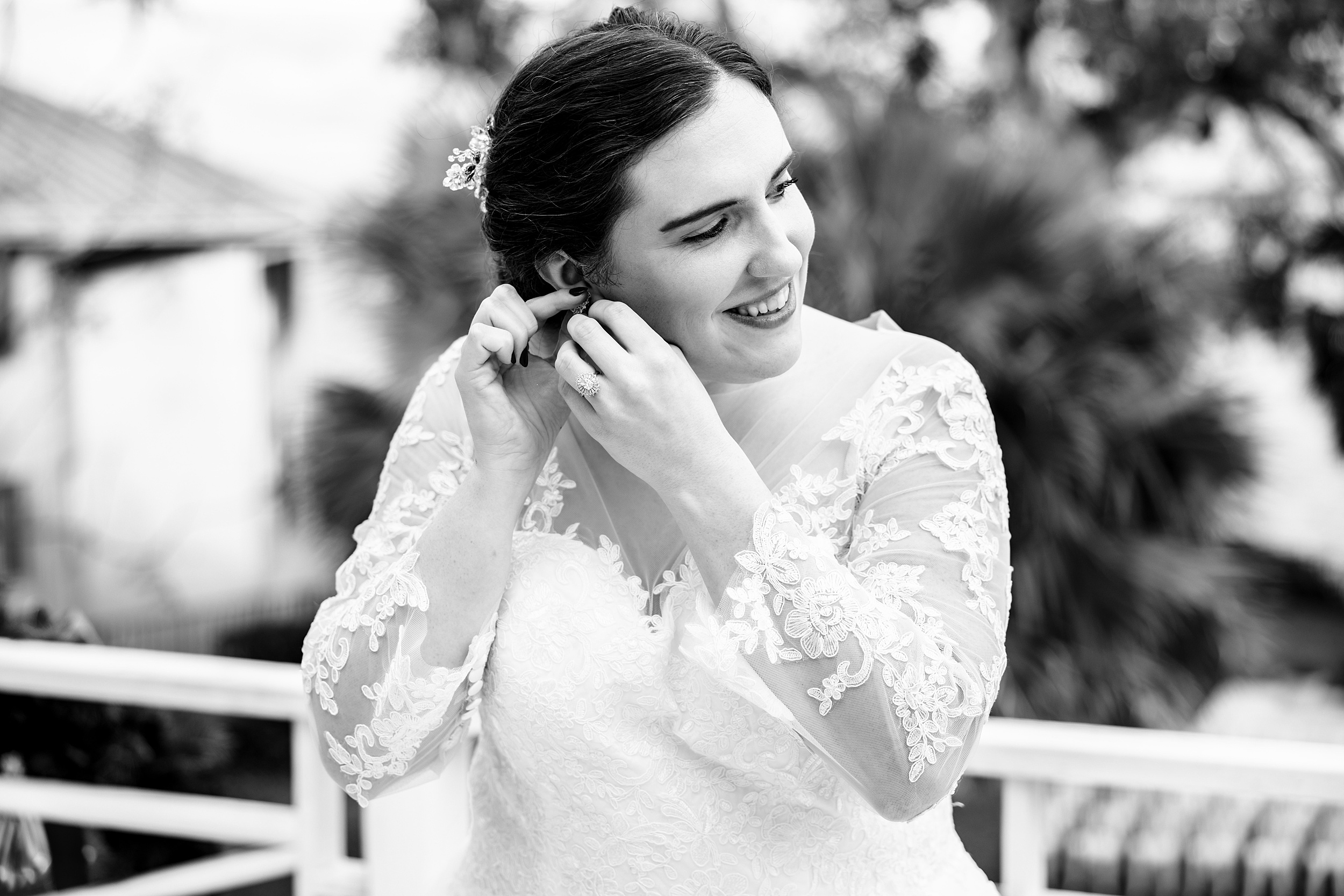 A happy bride puts on her earring while standing in a lace dress at her twin oak garden wedding